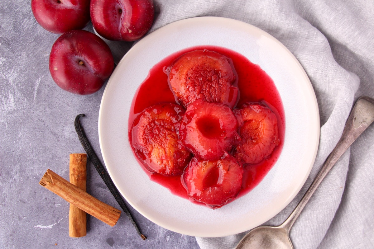 Stewed plums with a vintage serving spoon, fresh plums, cinnamon stick and vanilla pod on the side.