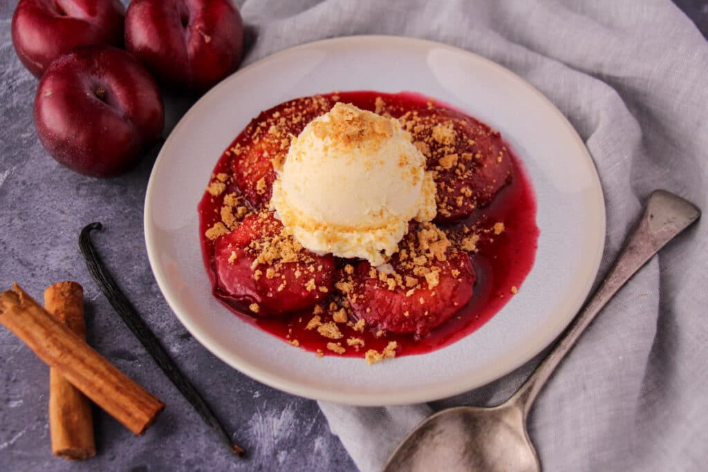 Stewed plums with a scoop of vanilla ice cream, biscotti crumb, a vintage serving spoon, fresh plums, cinnamon stick and vanilla pod on the side.