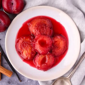Close up stewed plums with a vintage serving spoon, fresh plums, cinnamon stick and vanilla pod on the side.