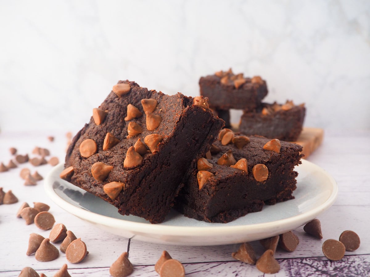 Two eggless brownies on a plate, with a stack of eggless brownies in the background, and chocolate chips scattered around.