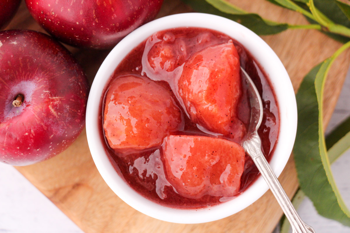 Plum compote in a serving bowl with a vintage spoon, fresh plums and leave on the side.