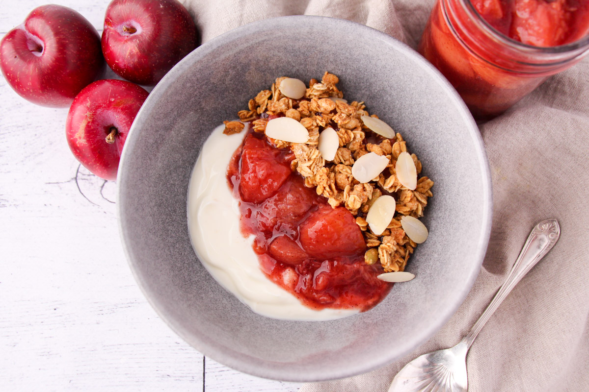 Serving suggestion of plum compote with vanilla yogurt and almond granola, with jar of plum compote, fresh plums and spoon on the side.