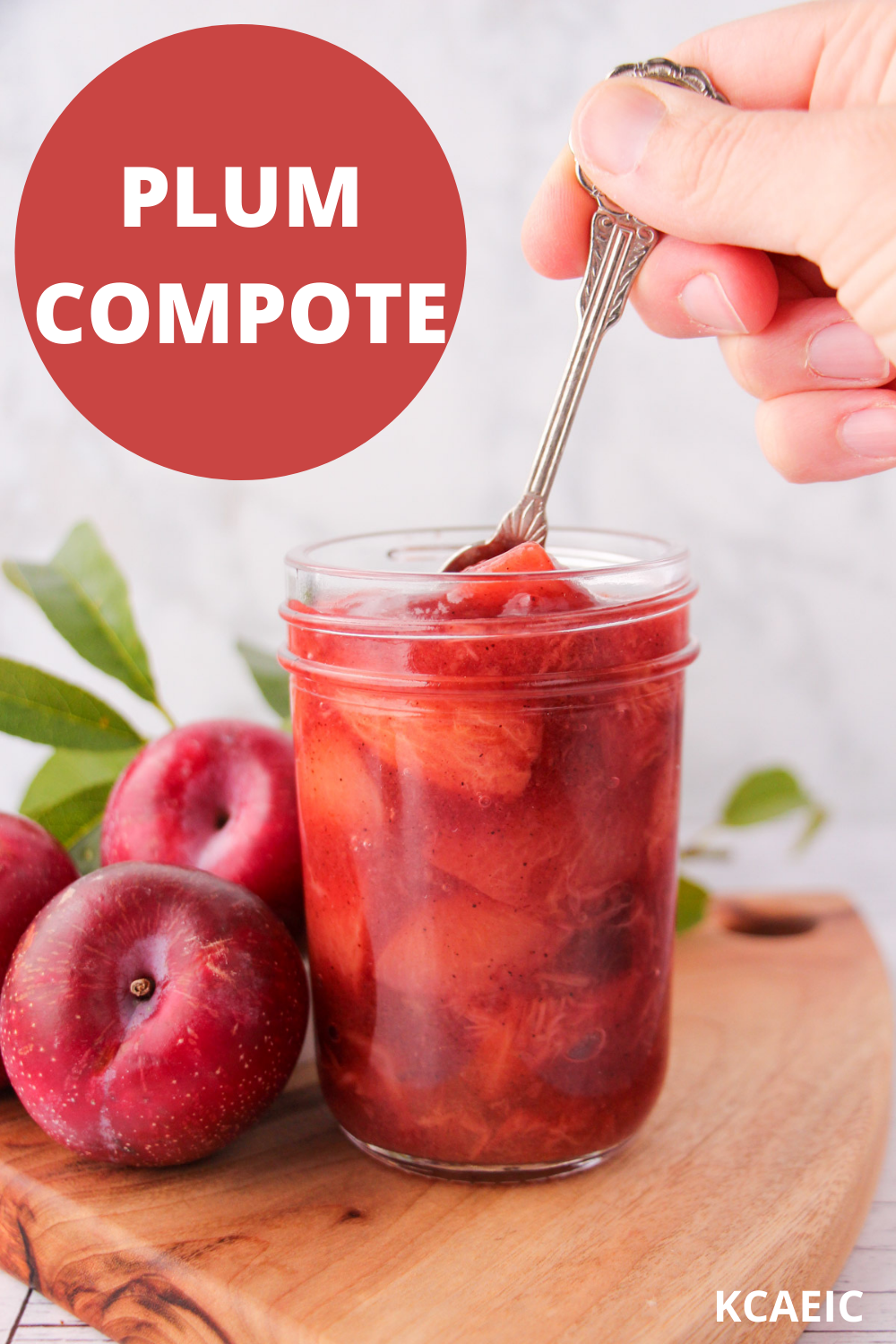 Mason jar of plum compote with a spoon being held over it, fresh plums and leave on the side and text overlay, plum compote, KCAEIC.