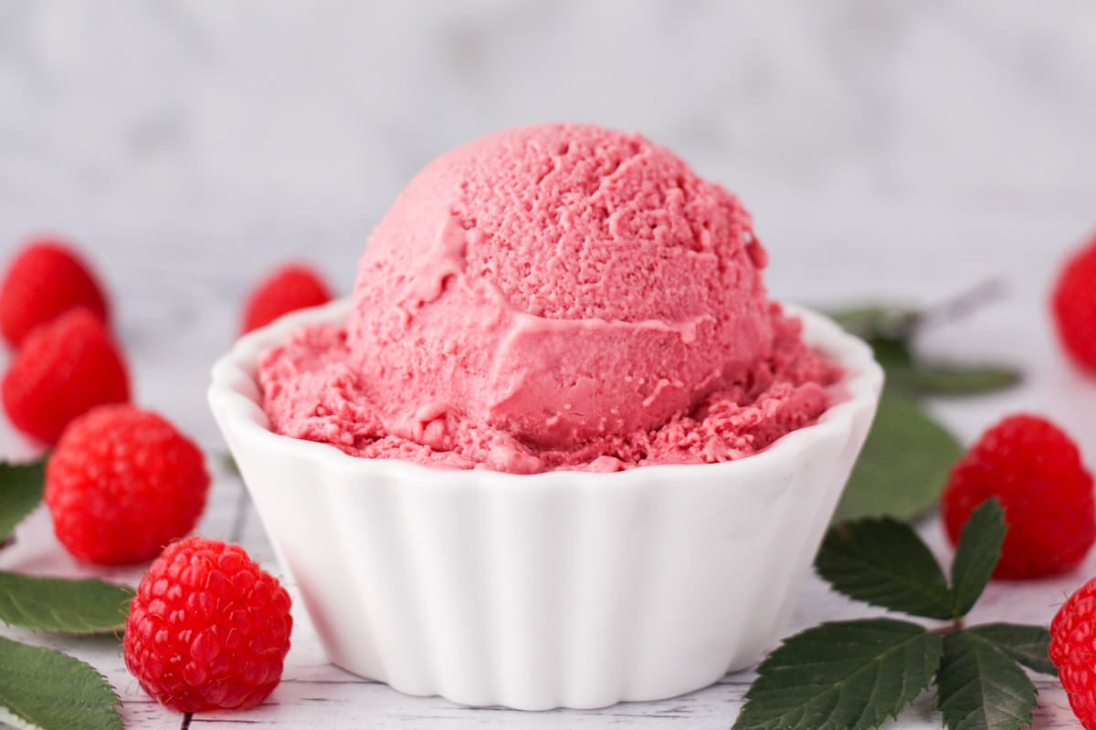 Scoop of raspberry ice cream in a bowl, with fresh raspberries and leaves on the side.