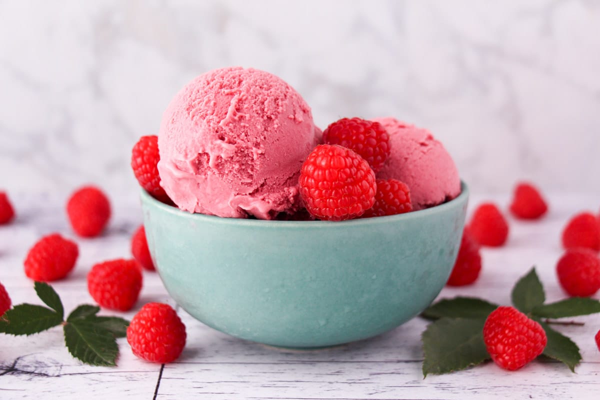 Scoop of raspberry ice cream in a bowl, with fresh raspberries and leaves on the side.