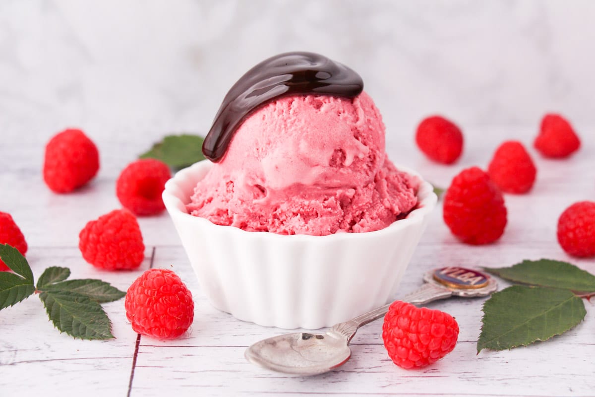 Scoop of raspberry ice cream with a drizzle of chocolate sauce in a bowl, with fresh raspberries and leaves on the side and a vintage spoon.