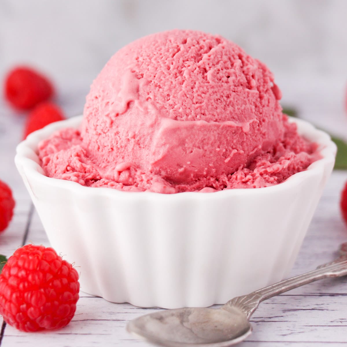 Close up of scoop of raspberry ice cream in a bowl, with fresh raspberries on the side and a vintage spoon.