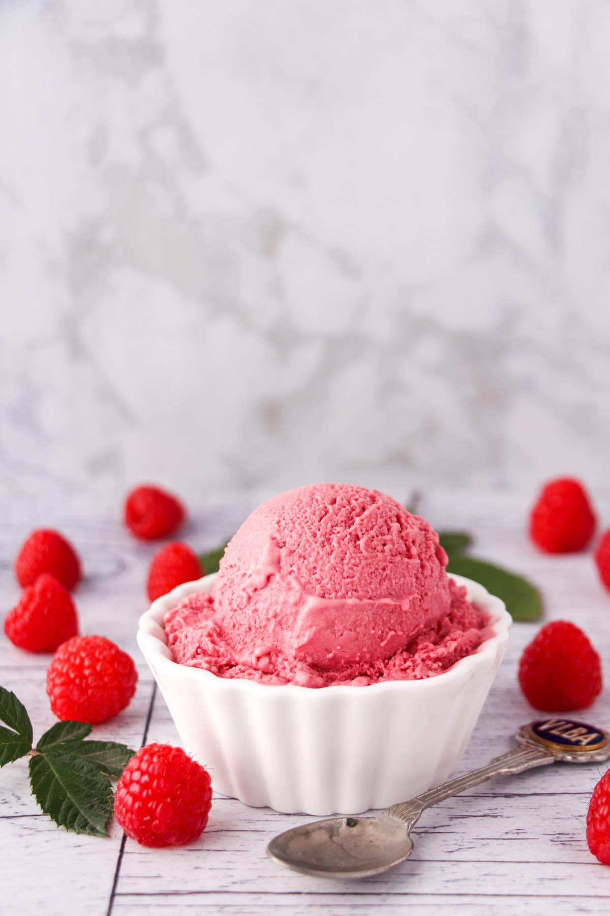 Scoop of raspberry ice cream in a bowl, with fresh raspberries and leaves on the side and a vintage spoon.