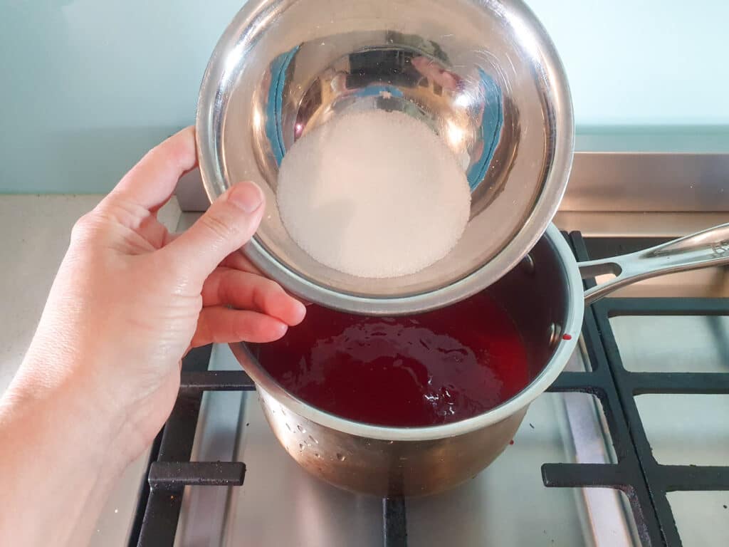 Adding sugar to strained raspberries in pot on stove.