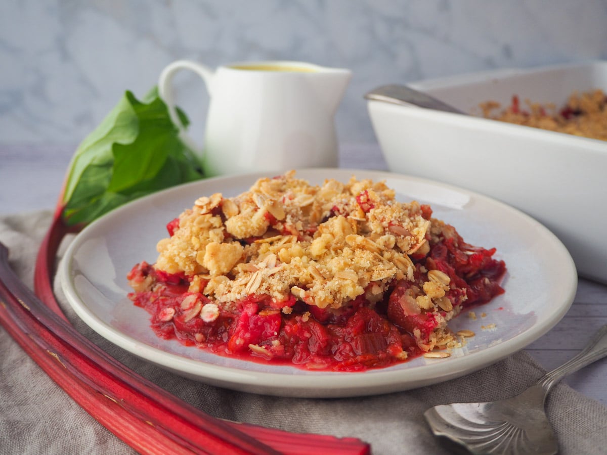 Serving rhubarb crumble with fresh rhubarb, vintage spoon, jug of custard and baking dish of crumble on the side.