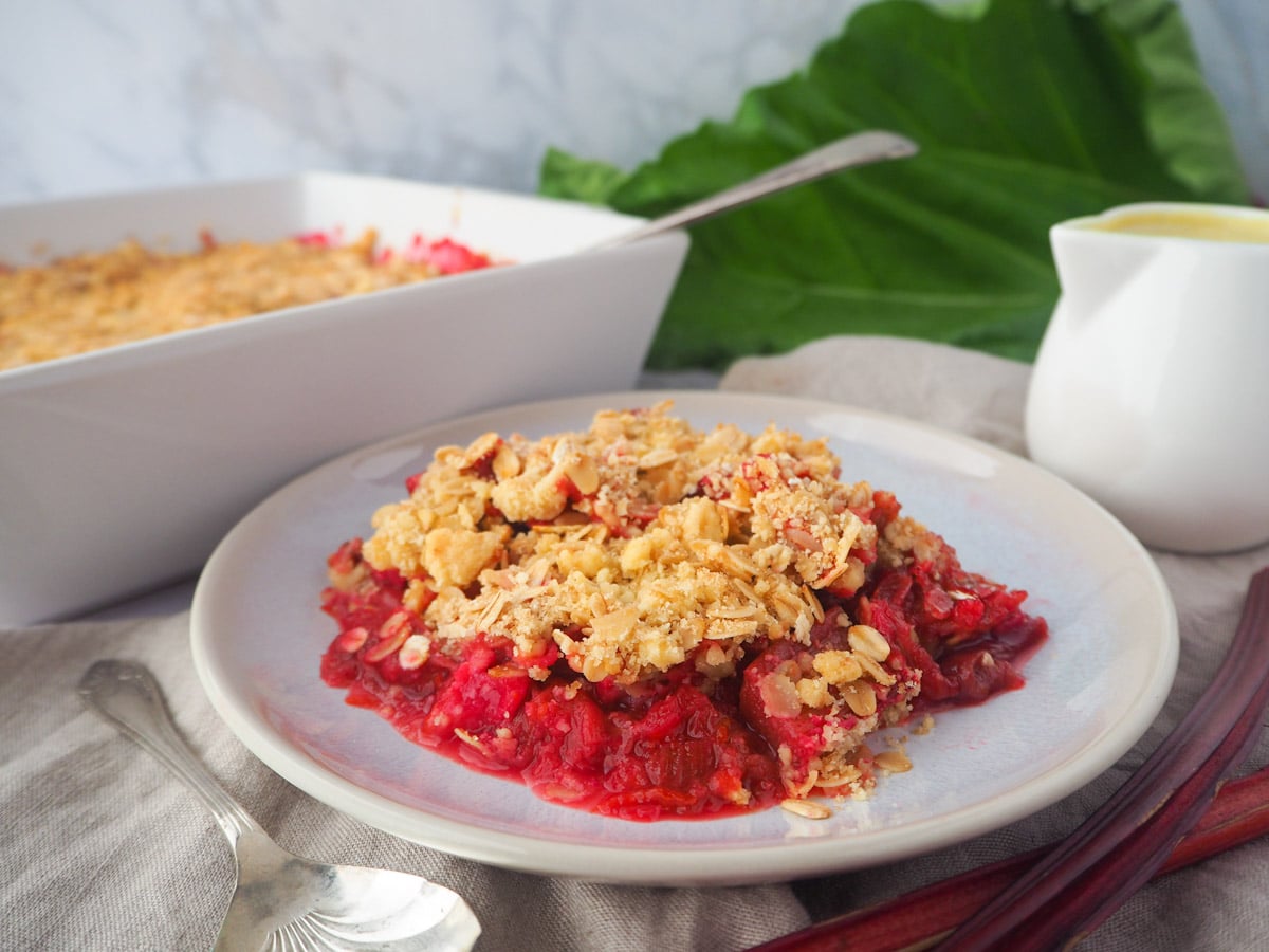 serving rhubarb crumble with custard and fresh rhubarb, baking dish of crumble and jug of custard on the side.