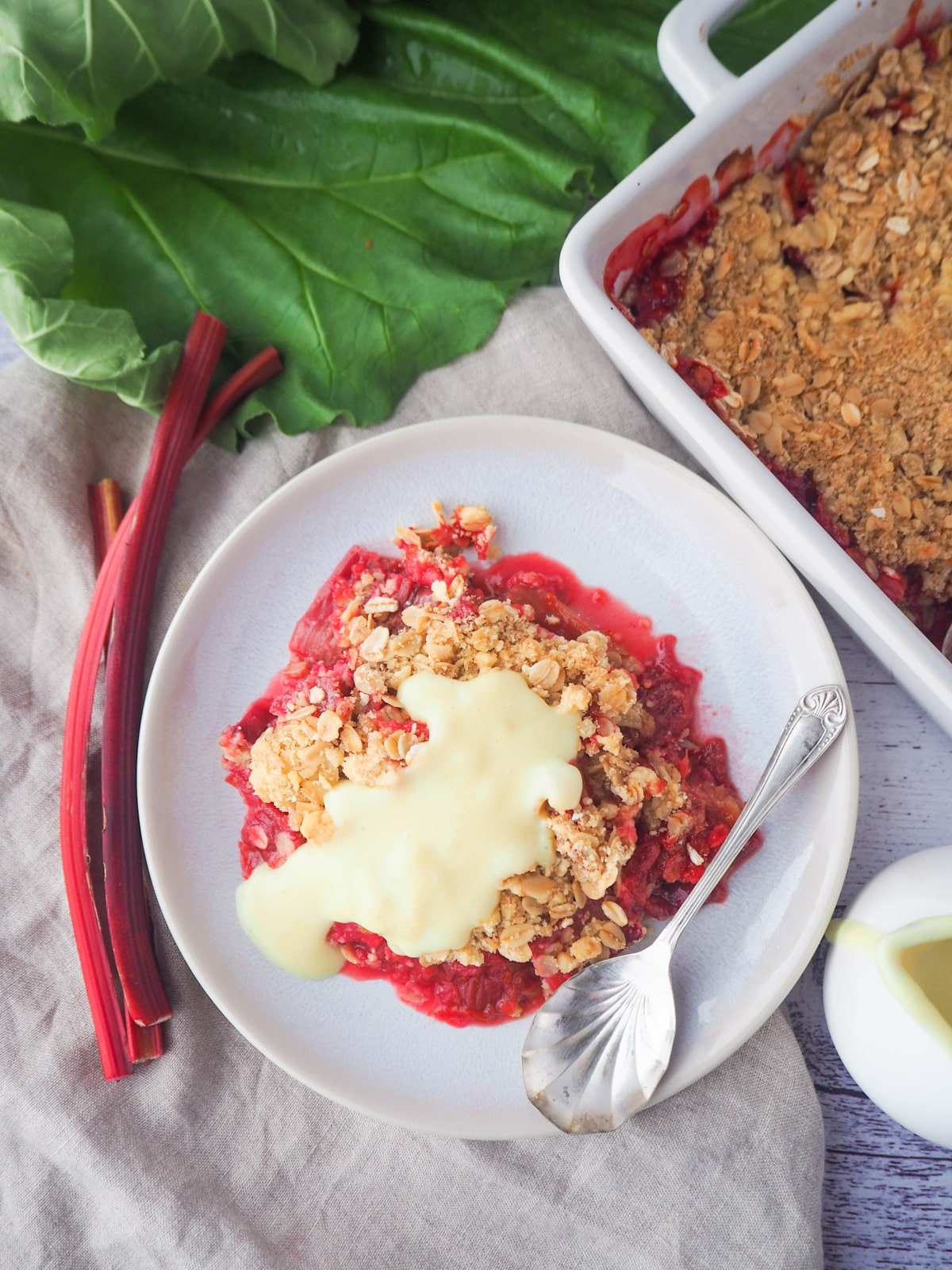 Top down view of serving rhubarb crumble with custard and fresh rhubarb, baking dish of crumble and jug of custard on the side.