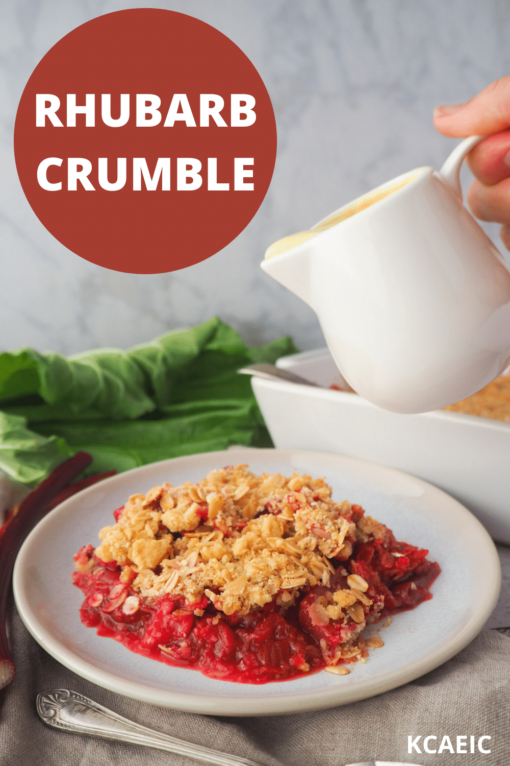 Serving of rhubarb crumble with jug of custard, with fresh rhubarb and baking dish in the background, with text overlay, rhubarb crumble and KCAEIC.