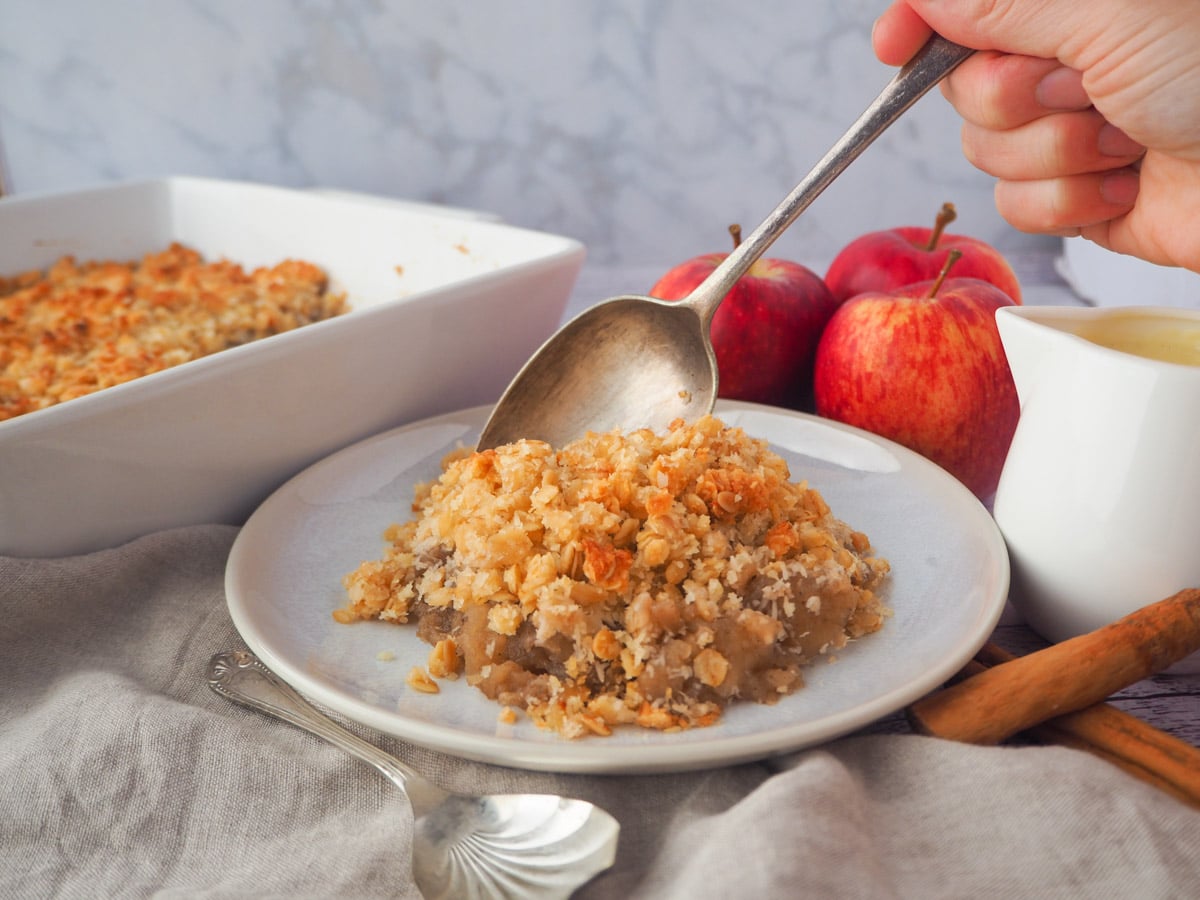 Serving vegan apple crumble with a vintage spoon, with fresh apples, jug of vegan custard and baking dish in the background.