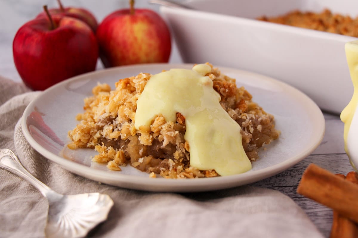 Vegan apple crumble with vegan custard, with fresh apples and baking dish in the background.