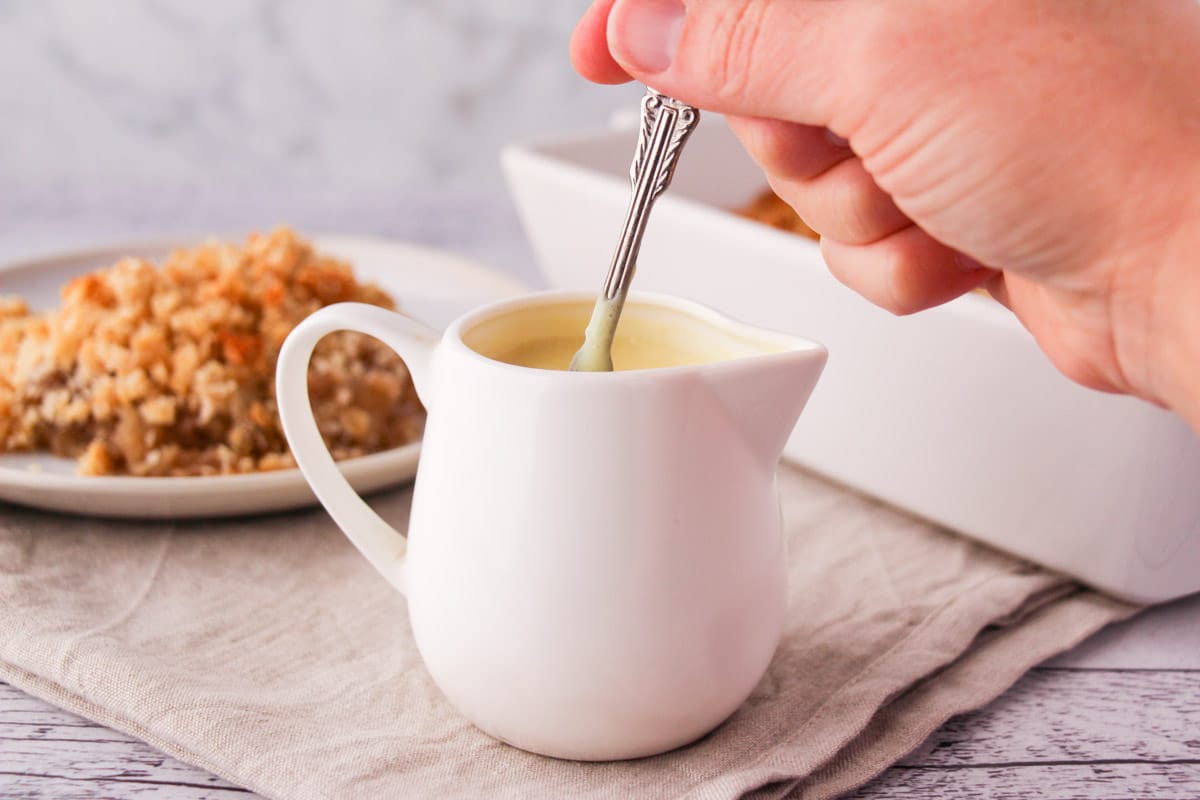 Spooning out vegan custard from a jug with a vintage spoon, with vegan apple crumble in the background.