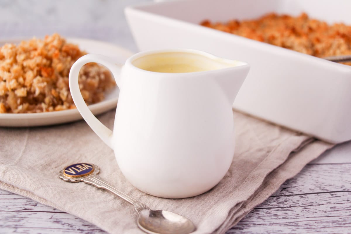 Jug of vegan custard with a vintage spoon and vegan apple crumble in the background.