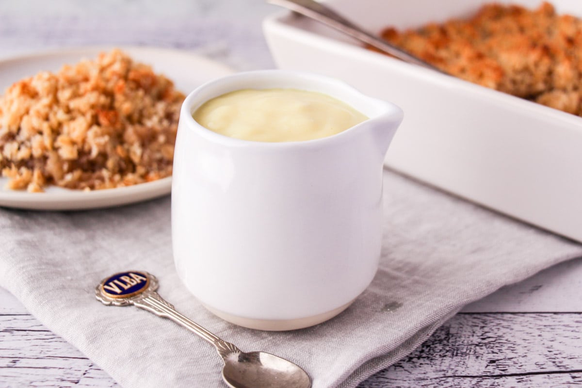 Jug of vegan custard with a vintage spoon and vegan apple crumble in the background.