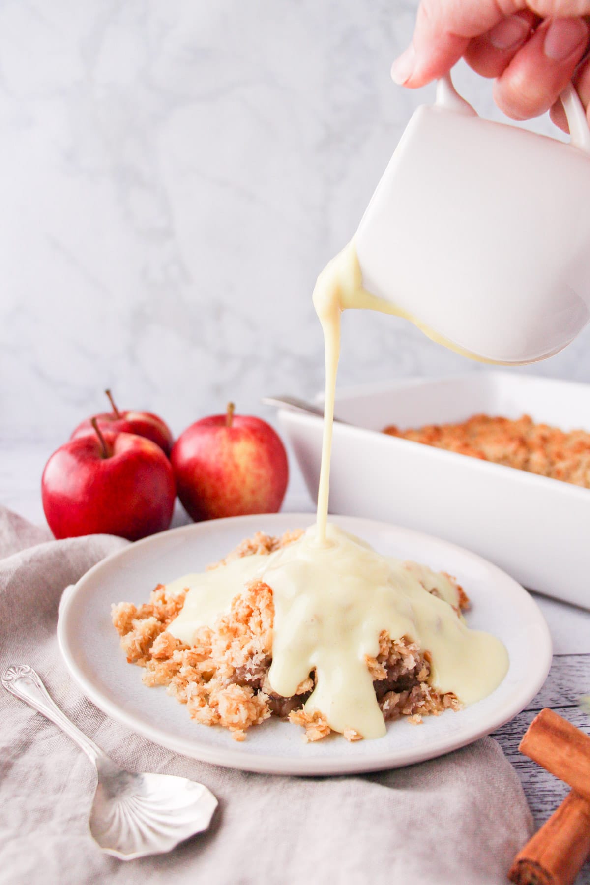 Vegan custard being poured over a vegan apple crumble, with apples and baking dish in background.