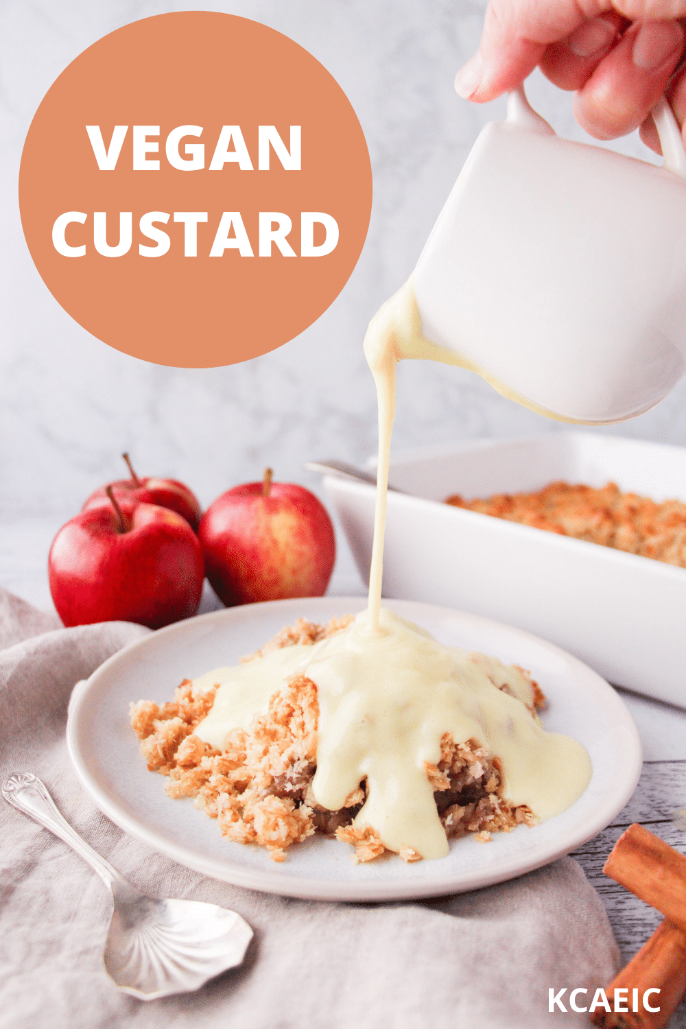 Vegan custard being poured over a vegan apple crumble, with apples and baking dish in background and text over lay, vegan custard and KCAEIC.