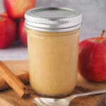 Close up jar of apple curd to spoon, fresh apples and cinnamon sticks.
