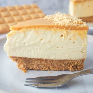 Close up slice of caramilk cheesecake on a plate with a fork with rest of cake and caramilk chocolate in the background.