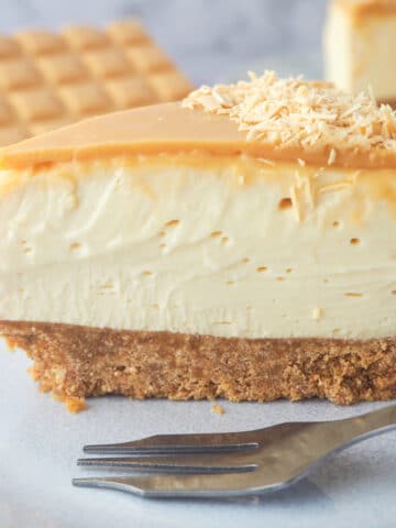 Close up slice of caramilk cheesecake on a plate with a fork with rest of cake and caramilk chocolate in the background.