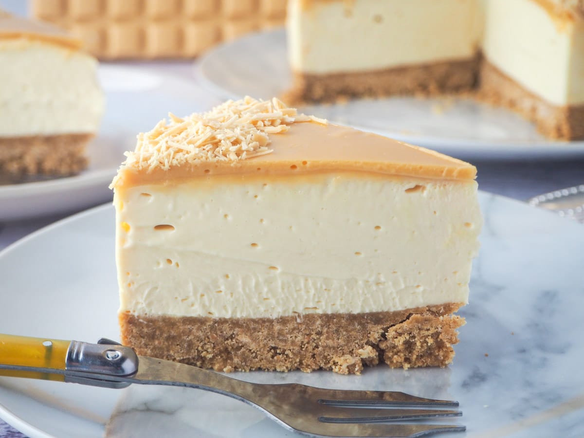 Slice of caramilk cheesecake on a plate with a fork, rest of cake, extra slice of cake and caramilk chocolate in the background.