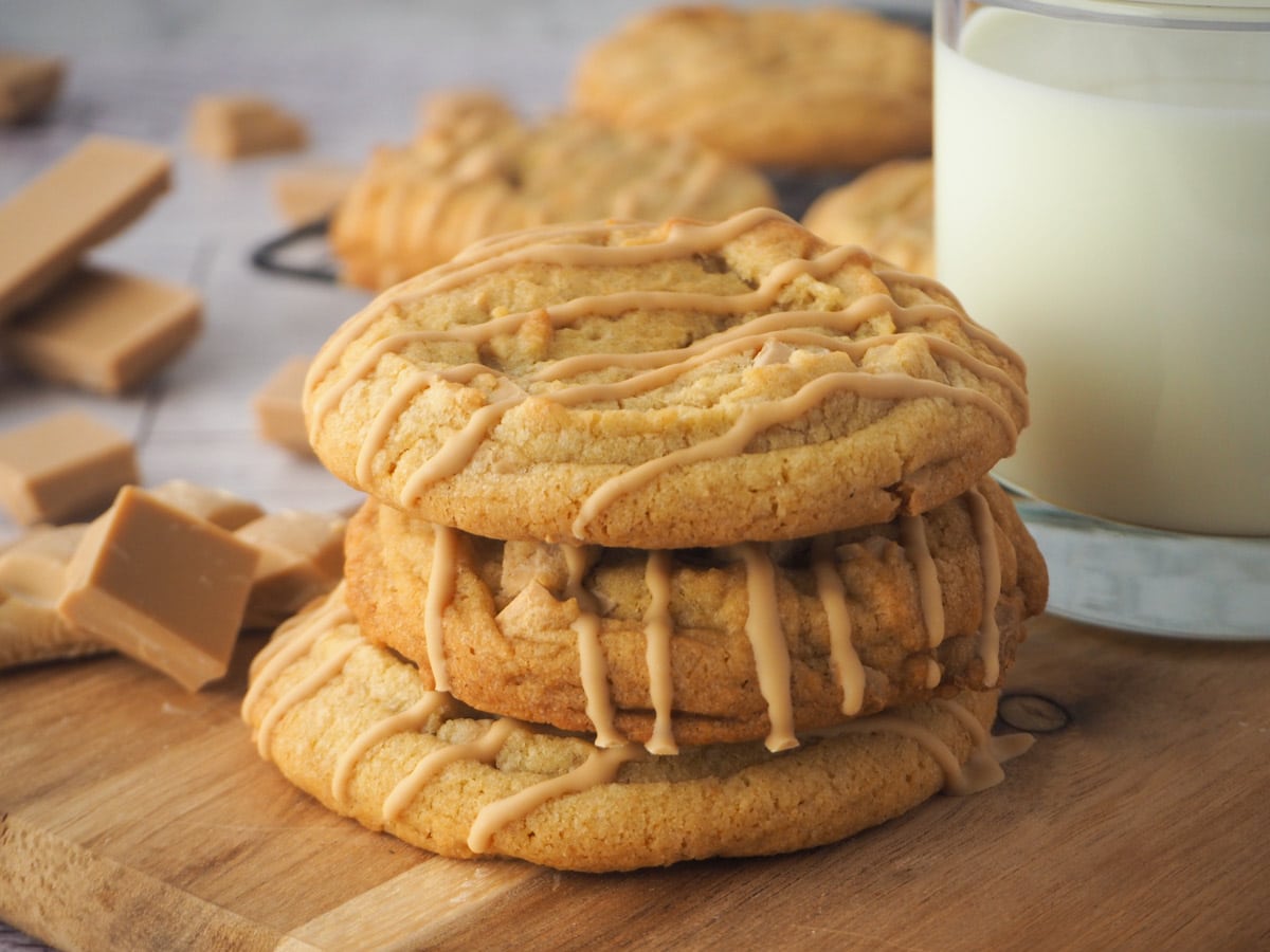 Stack of caramilk cookies o a board with a glass of milk and caramilk chocolate and more cookies in the background.