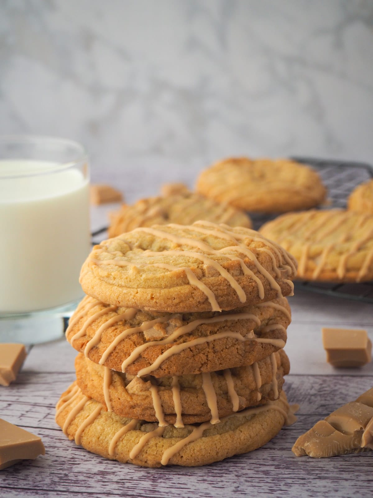 Stack of caramilk cookies o a board with a glass of milk and caramilk chocolate and more cookies in the background.