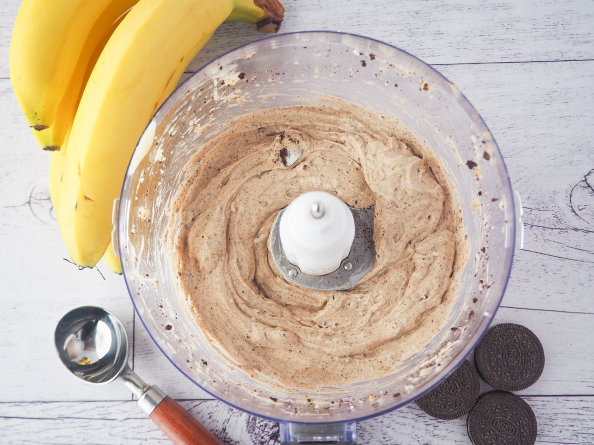 Top down view of open food processor with cookies and cream nice cream, with bananas, Oreos and ice cream scoop on the side.