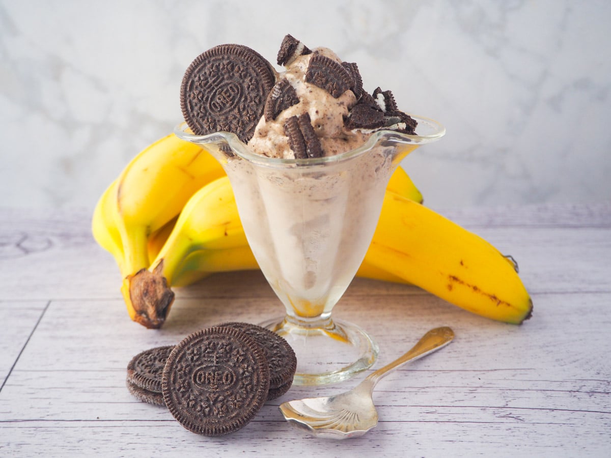 Serve of cookies and cream nice cream in a sundae glass, with pieces of Oreos, bananas and whole Oreos in the background.