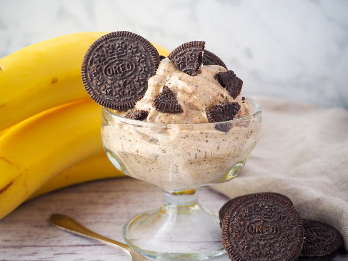 Serve of cookies and cream nice cream in a bowl, with pieces of Oreos, bananas and whole Oreos in the background.