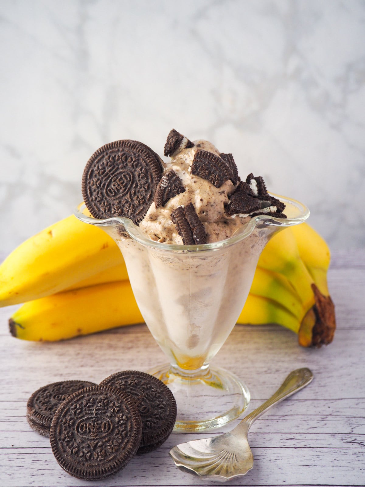 Serve of cookies and cream nice cream in a sundae glass, with pieces of Oreos, bananas and whole Oreos in the background.