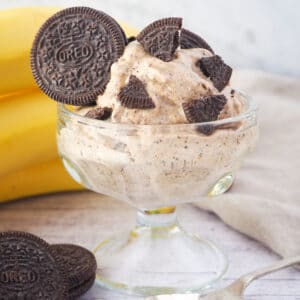 Close up Serve of cookies and cream nice cream in a bowl, with pieces of Oreos, bananas and whole Oreos in the background.