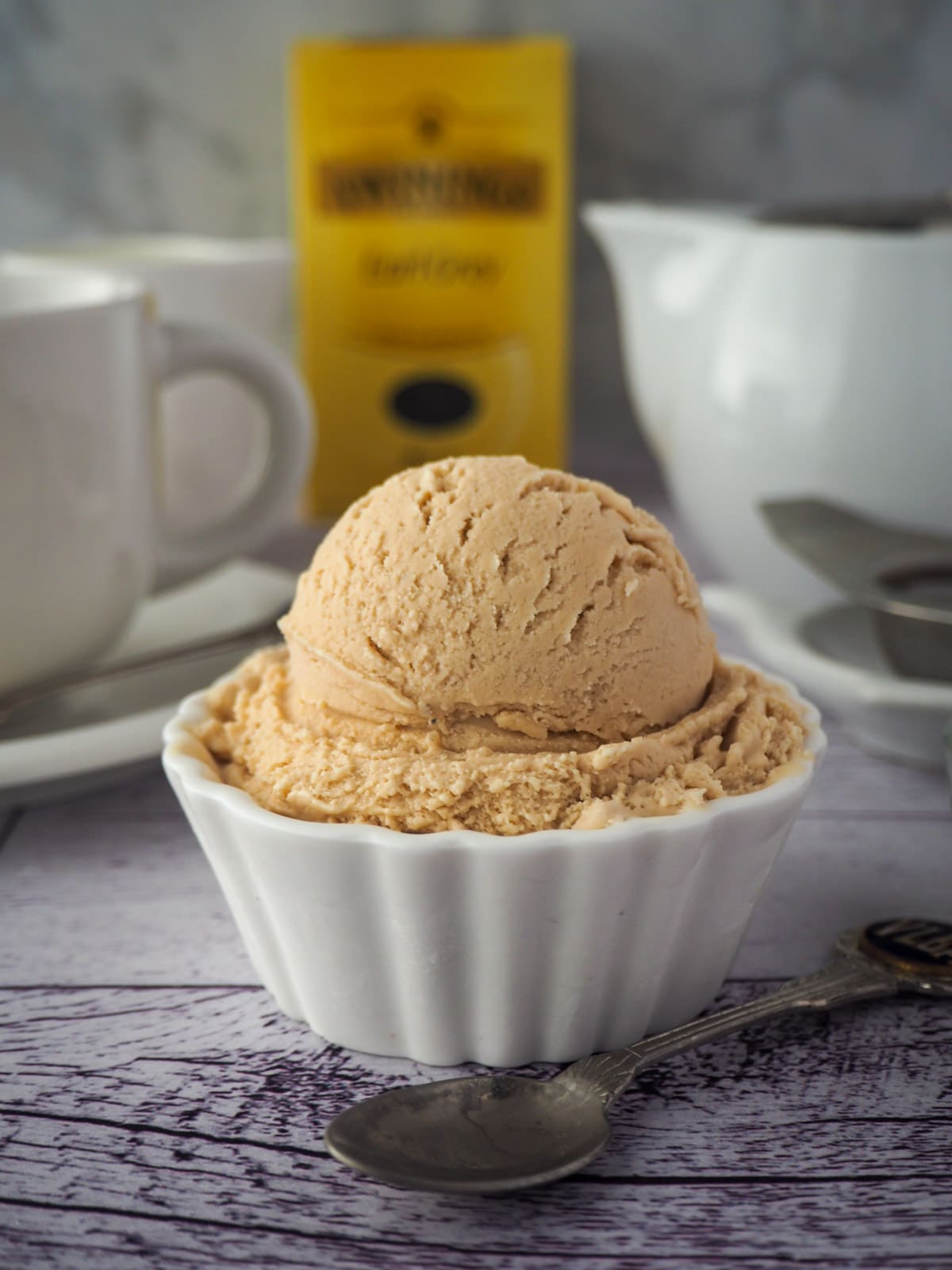 Scoop of earl grey ice cream with spoon and cut of tea, tea pot and box of tea in the background.