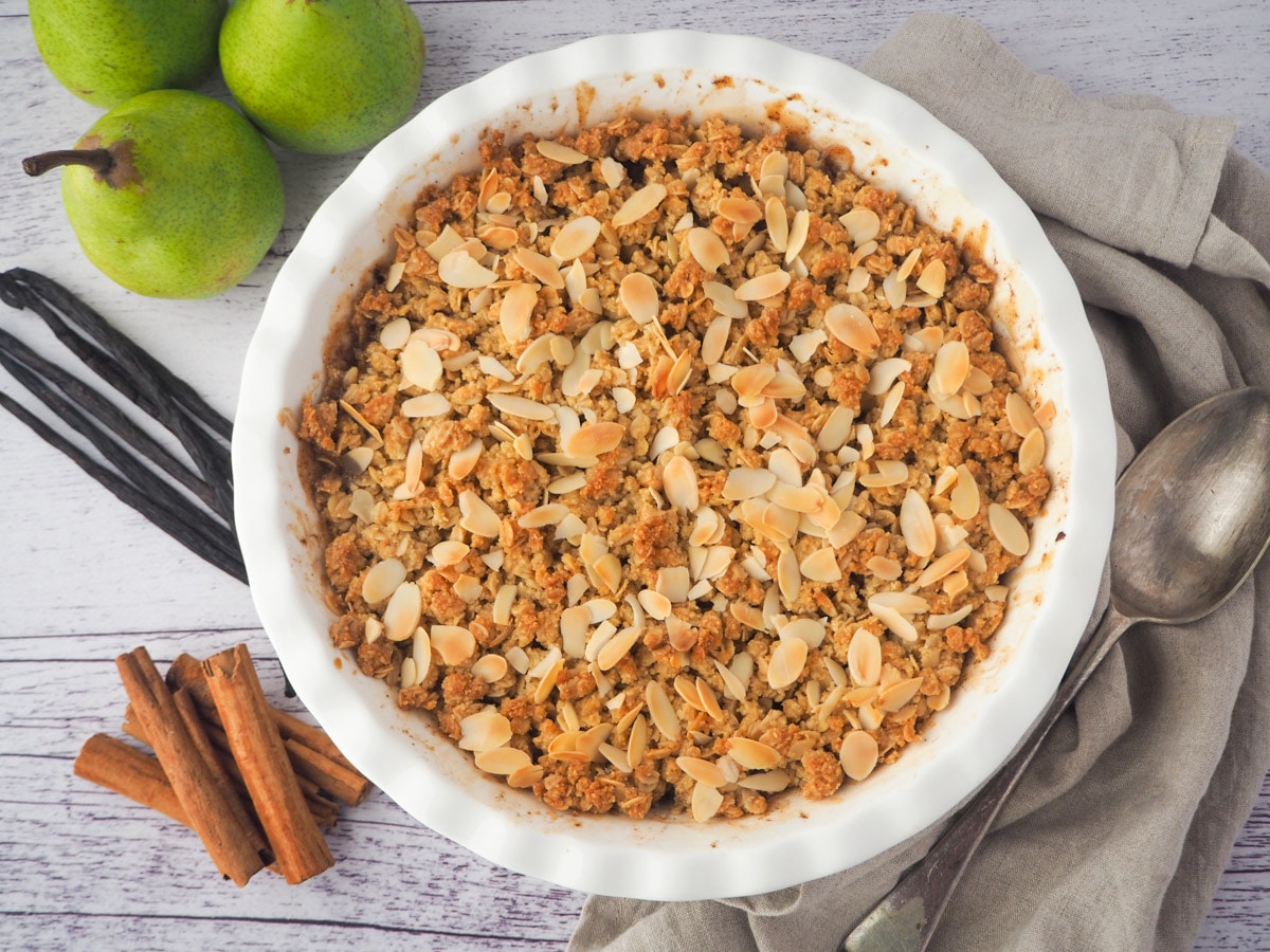 Pear crisp in baking dish with serving spoon, fresh pears, cinnamon sticks and vanilla pods on the side.