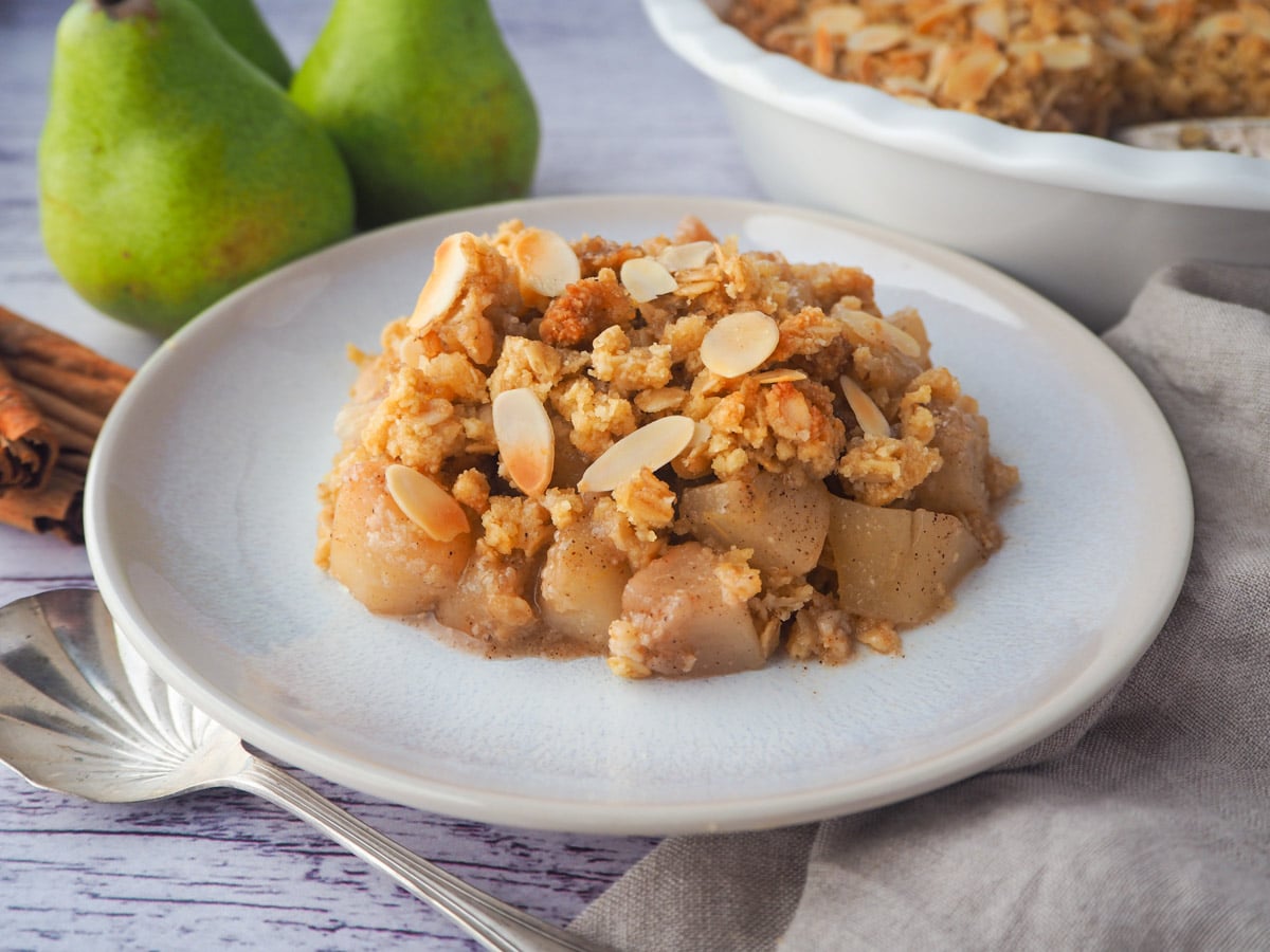 Serving of pear crumble with spoon, fresh pears, cinnamon sticks and baking dish of crumble in the background,