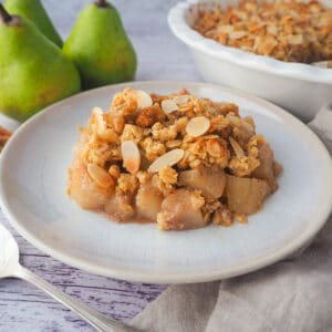 Close up Serving of pear crumble with spoon, fresh pears, jug of custard and baking dish of crumble in the background.