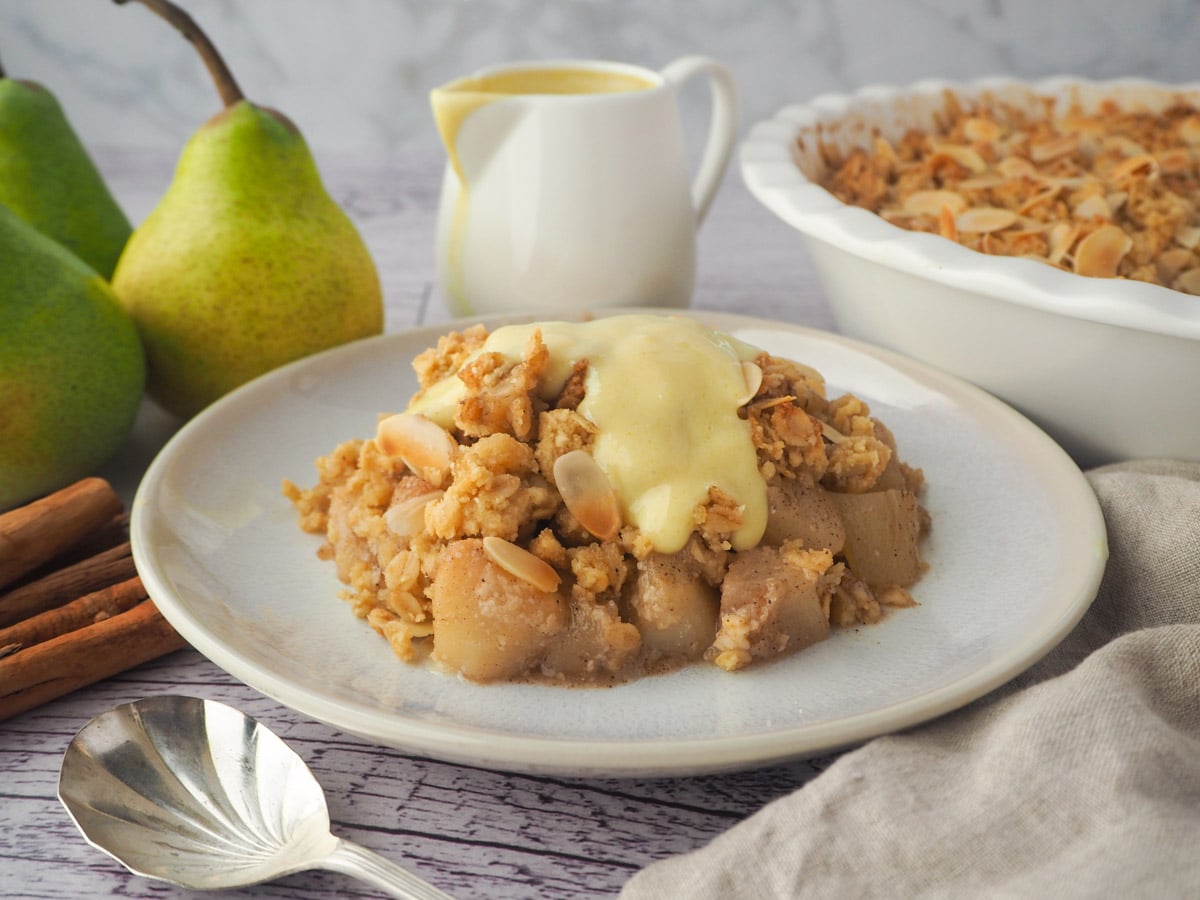 Serving of pear crumble with custard with spoon, fresh pears, cinnamon sticks, jug of custard and baking dish of crumble in the background,