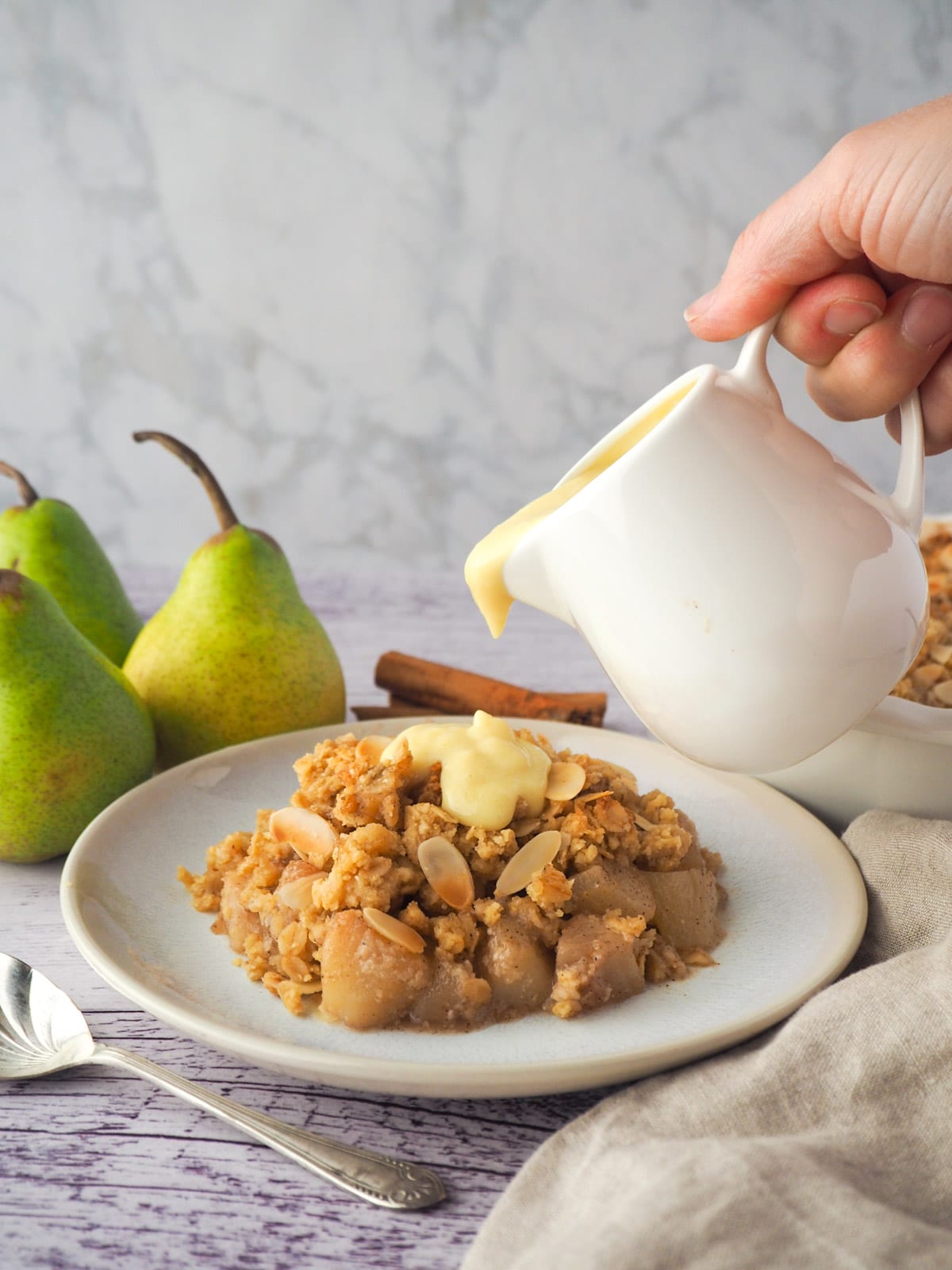 Pear crumble 18 Serving of pear crumble with custard being poured over the top, with fresh pears, cinnamon stick and crumble in baking dish in background.