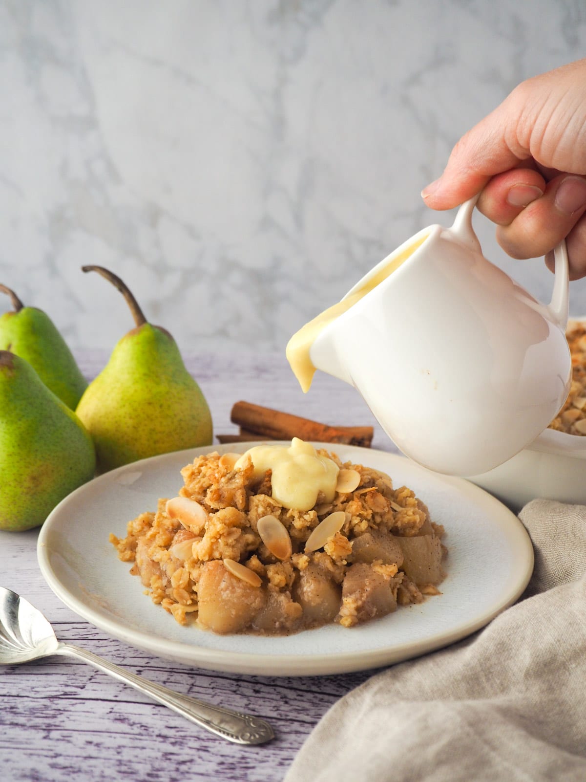 Serving of pear crumble with custard being poured over the top, with fresh pears, cinnamon stick and crumble in baking dish in background.