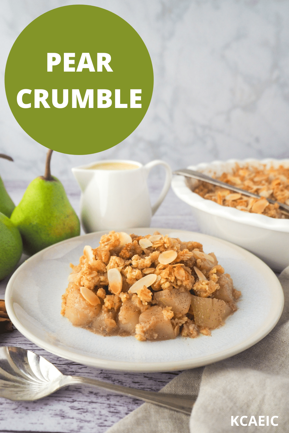 Serving of pear crumble with spoon, fresh pears, jug of custard and baking dish of crumble in the background, with text overlay, pear crumble and KCAEIC.