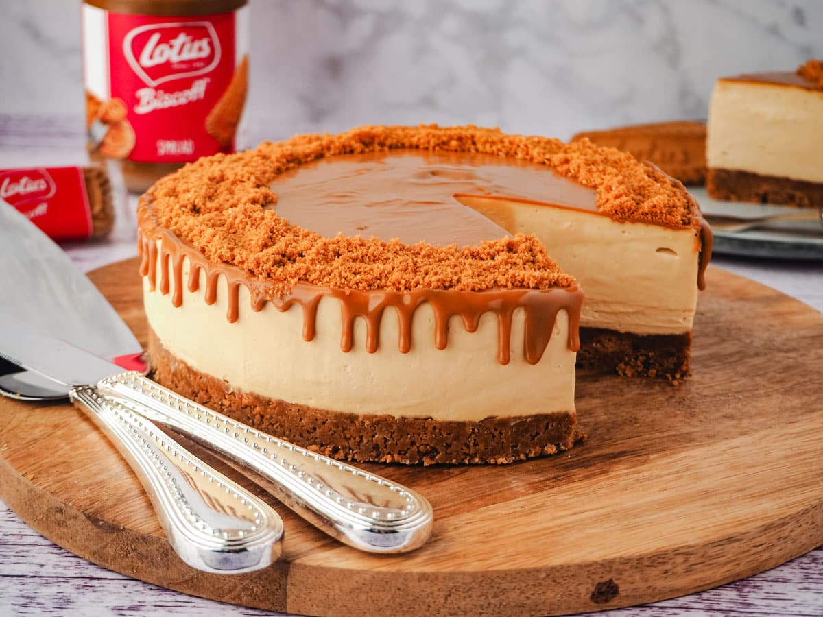 Cheesecake on a serving board with serving wear, jar of Biscoff and Biscoff cookies and slice of cake in background.