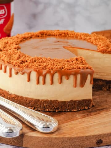 Cheesecake on a serving board with serving wear, jar of Biscoff and Biscoff cookies and slice of cake in background.