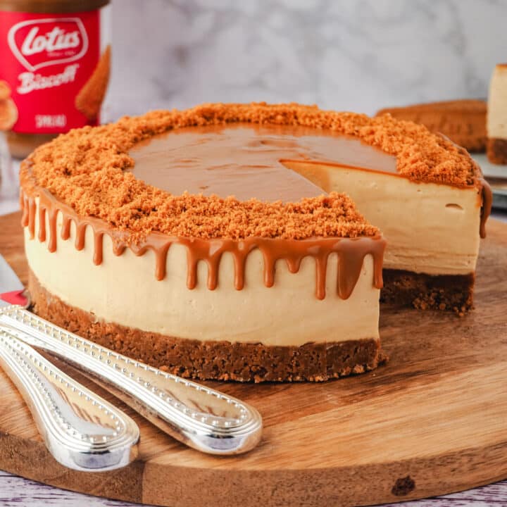 Cheesecake on a serving board with serving wear, jar of Biscoff and Biscoff cookies and slice of cake in background.