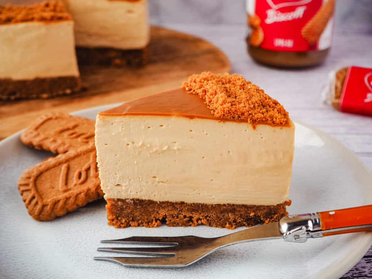 Slice of cheesecake on a plate with fork and Biscoff biscuits, with rest of cake, jar of Biscoff and Biscoff biscuits in the background.