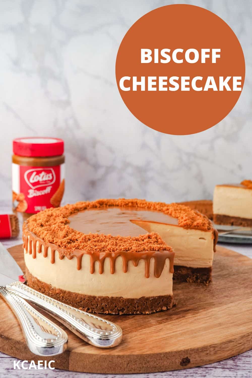 Cheesecake on a serving board with serving wear, jar of Biscoff and Biscoff cookies and slice of cake in background, with text overlay, Biscoff Cheesecake and KCAEIC.