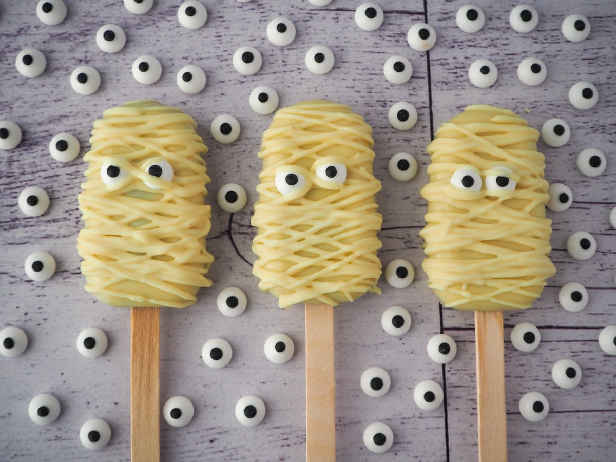 Row of three mummy cakesicles, surrounded by candy eyes.