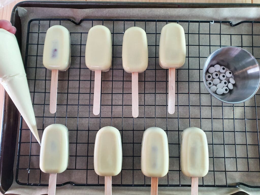 Halloween cakesicles 31 Mummy cakesicles laid out ready to decorate, with candy eyes and piping bag of melted chocolate.